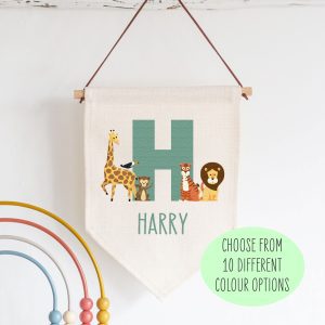 Personalised jungle safari linen flag for a nursery. The flag is a pale beige colour with a wooden dowel at the top, hanging from a brown string. A large, turquoise letter "H" is in the centre, with a cartoon giraffe, monkey, tiger, and lion on either side of it. Below the animals, the name "Harry" is written in a matching turquoise font. The flag is a perfect newborn baby boy or girl nursery gift. It is a lovely birthday gift for a son or daughter's bedroom decor.