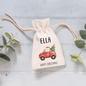 A linen-style drawstring gift bag with a red vintage car and a Christmas tree on top, driven by Santa. The customisable name "ELLA" is printed above the design, with "HAPPY CHRISTMAS" below. The bag sits on a light wooden surface next to green leaves. This personalised Christmas pouch is perfect for gift-giving and stocking fillers, adding a festive touch to any present. It's an ideal choice for Christmas Eve boxes and unique holiday gift-wrapping.