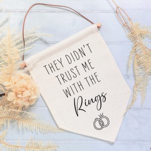 A small, natural-coloured linen flag hangs from a wooden dowel with a faux suede cord. The flag is printed with the funny wedding quote "They Didn't Trust Me With The Rings" and an icon of two interlocking wedding rings. The flag is for a best man, page boy or groomsman to carry down the aisle.