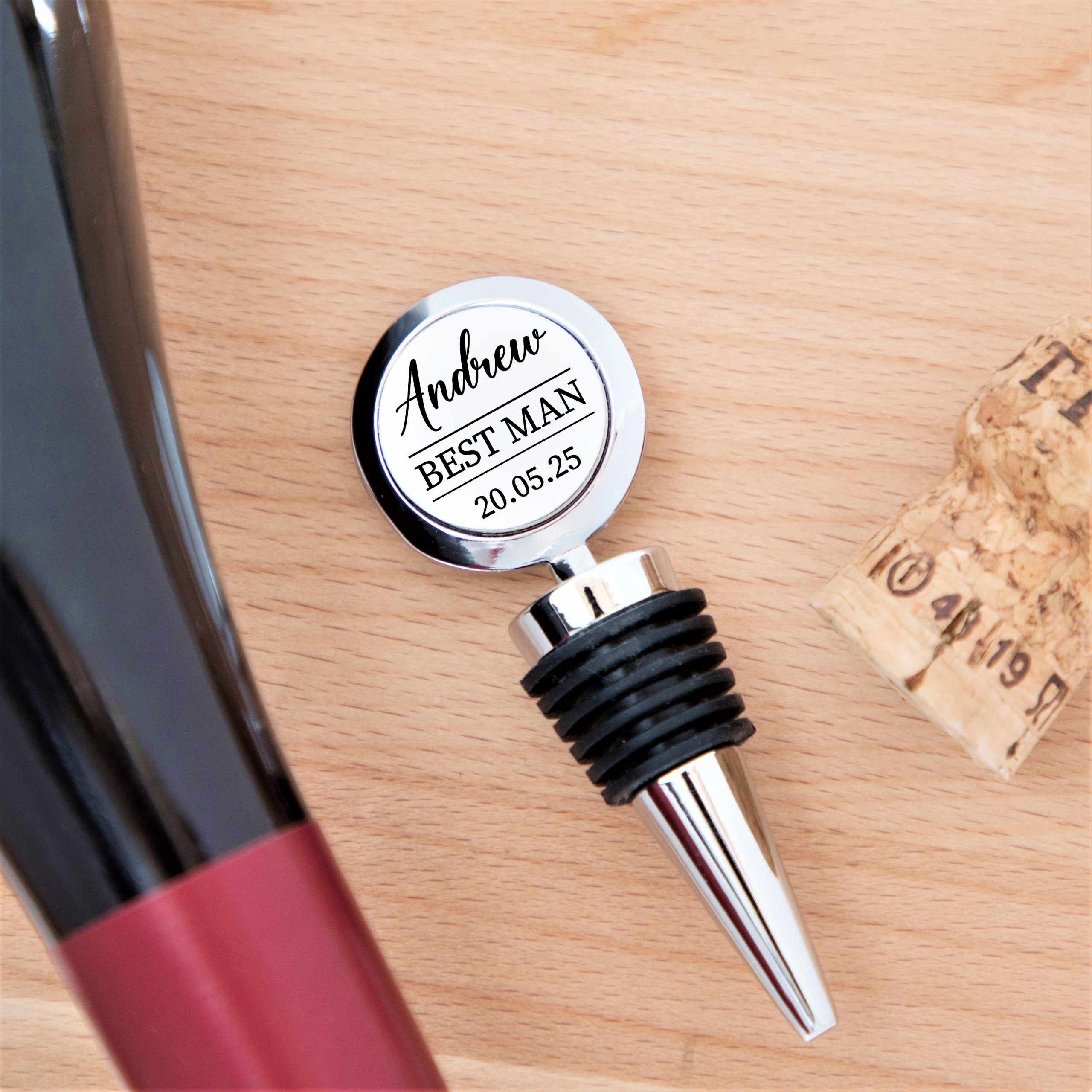 A personalised metal bottle stopper is shown on a light wooden surface. The round, chrome-finished top has a white insert with black text. The customisable text reads "Paul", "GROOMSMAN", and the date "12.04.26". A black rubber section is visible below the top, and a wine bottle cork is partially shown on the right. This unique gift is perfect for a groom, best man, or groomsman, maid of honour or bridesmaid.