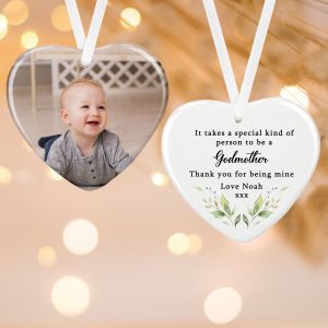 A close-up shot of a personalised heart-shaped ceramic decoration. On the left is a heart with a picture of a smiling baby. On the right is a second heart, featuring text that reads "It takes a special kind of person to be a Godmother Thank you for being mine Love Noah xxx". The hearts are shown against a soft-focus background of fairy lights and are hanging from white ribbons. The product is a beautiful, personalised keepsake gift for a godmother.