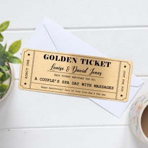 A rectangular golden metal voucher card designed like a "Golden Ticket" with personalized text. The top line reads "GOLDEN TICKET" in a large, bold font. Below that, in a script font, is "Louise & David Jones". Further down, it states "THIS TICKET ENTITLES YOU TO: A COUPLE'S SPA DAY WITH MASSAGES". At the bottom, there's a personalized message: "Happy Anniversary! Lots of love from Mum & Dad xxx". The card has a decorative border with stars and "ADMIT ONE" vertically on the left side, and "JUST FOR YOU" vertically on the right side. The ticket is shown resting on a white envelope, with a green plant in a white pot on the left and a part of a patterned mug on the right, all on a light wooden surface.