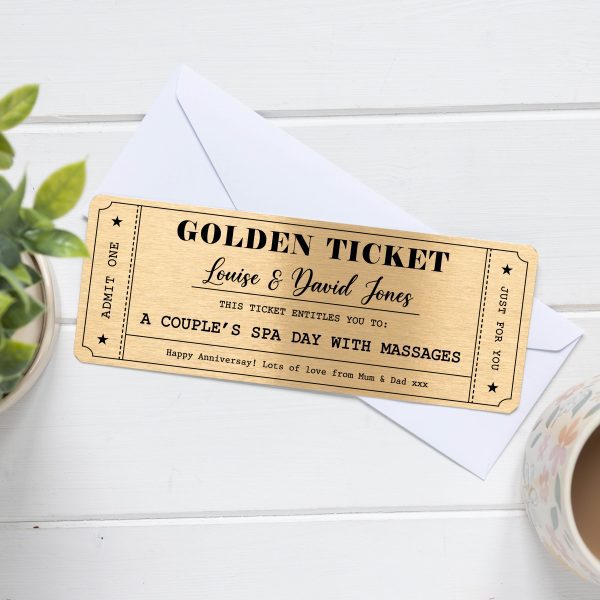 A rectangular golden metal voucher card designed like a "Golden Ticket" with personalized text. The top line reads "GOLDEN TICKET" in a large, bold font. Below that, in a script font, is "Louise & David Jones". Further down, it states "THIS TICKET ENTITLES YOU TO: A COUPLE'S SPA DAY WITH MASSAGES". At the bottom, there's a personalized message: "Happy Anniversary! Lots of love from Mum & Dad xxx". The card has a decorative border with stars and "ADMIT ONE" vertically on the left side, and "JUST FOR YOU" vertically on the right side. The ticket is shown resting on a white envelope, with a green plant in a white pot on the left and a part of a patterned mug on the right, all on a light wooden surface.