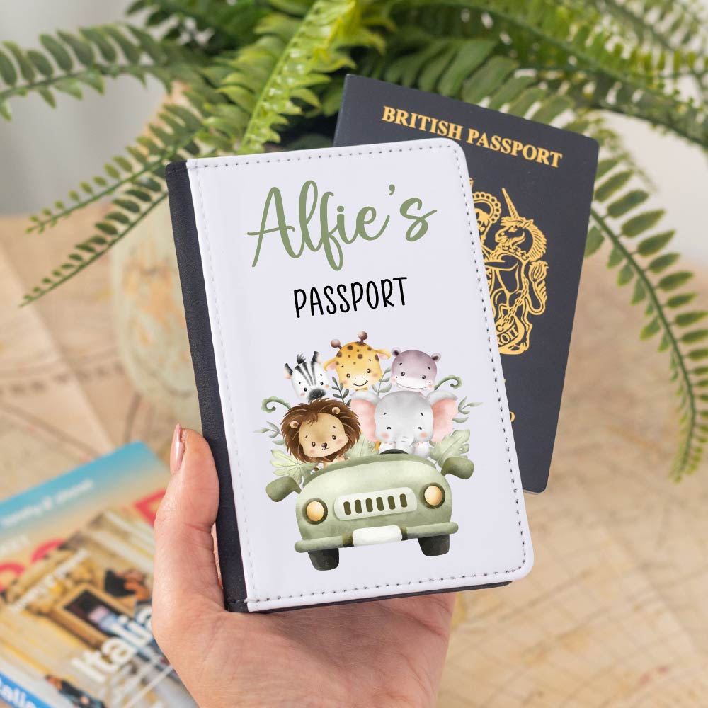 A child's white faux leather passport holder, personalized with "Alfie's PASSPORT" in green and black text. It features a cute safari scene with a lion, giraffe, zebra, hippopotamus, and elephant riding in a green jeep. A British passport is visible behind the holder. This custom travel wallet is perfect for a baby's first holiday, a unique gift for a boy or girl, or a christening present, offering a cute jungle animal design.