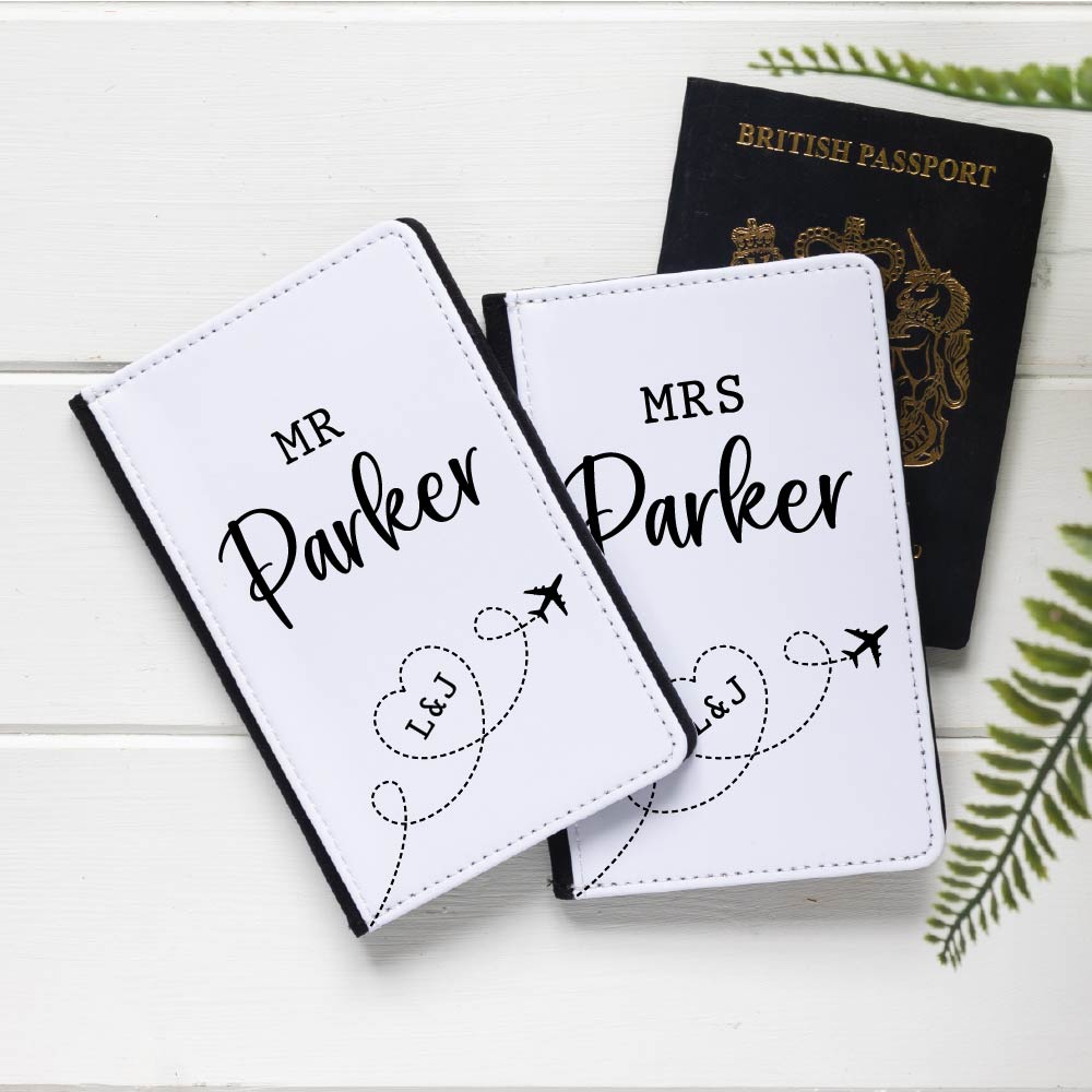 A pair of white faux leather passport holders for "Mr. Parker" and "Mrs. Parker" with custom initials "L & J" inside a heart, connected by dotted lines to an airplane icon. A British passport is partially visible behind them. Perfect personalized gift for newlyweds, anniversaries, honeymoons, or couples' travel.