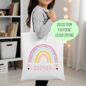 A young child holds a white personalised canvas tote bag featuring a pastel rainbow design and the customisable name "SOPHIA." The bag is a perfect gift for birthdays, back-to-school, or special occasions, and is suitable for children to use as a book bag or for carrying their belongings.