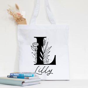 A close-up shot of a white fabric tote bag with a custom black floral design. A large customised letter 'L' is filled with delicate flowers, leaves, and buds. Below the initial, the customised name 'Lilly' is written in a graceful cursive font. The bag hangs against a white wall. The image is bright, clean, and highlights the personalised detail of the tote bag. Ideal gift for her, Mum, teacher or a bridesmaid.