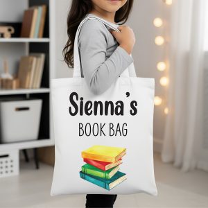 A young girl holds a white canvas book bag with the customisable name "Sienna's" and "BOOK BAG" printed above a stack of colourful rainbow-printed books. The durable, personalised tote is a perfect back-to-school or birthday gift for a book lover.