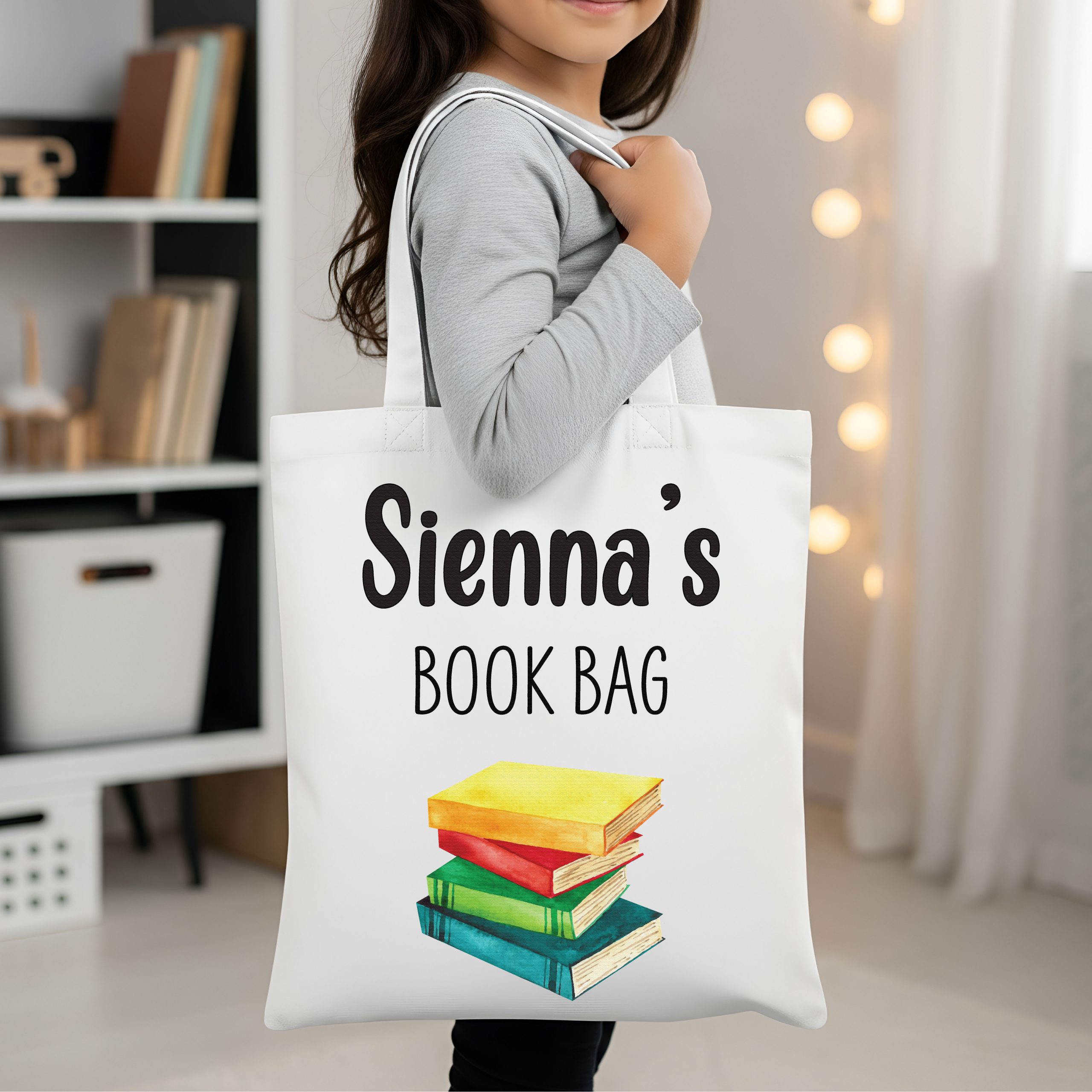 A young girl holds a white canvas book bag with the customisable name "Sienna's" and "BOOK BAG" printed above a stack of colourful rainbow-printed books. The durable, personalised tote is a perfect back-to-school or birthday gift for a book lover.