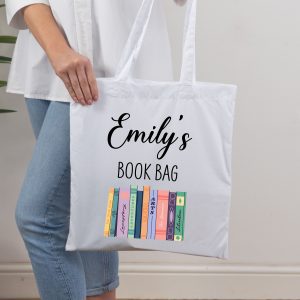 A white tote bag is held by a person wearing a white shirt and jeans. The bag has the customisable name "Emily's" in a black script font, with "BOOK BAG" in a simple black font underneath. Below this text is a colourful design of illustrated book spines with various titles. The bag is a personalised custom name fabric tote, ideal as a literary gift or bookworm present.