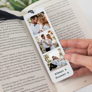 Personalised metal bookmark featuring three custom photo prints of a couple and the text "Always & Forever x". The bookmark has a black tassel and is shown holding the pages of a book. The background is a close-up of the book's pages, which have text on them. The bookmark is designed for couples, perfect for a wedding, anniversary, or Valentine's Day gift.