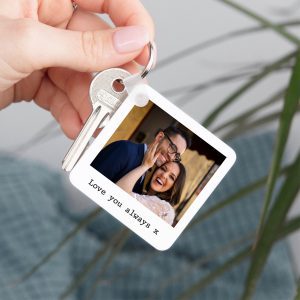 A personalised photo keyring featuring a couple's photo and a "Love you always x" message. The keyring is made of white plastic in a polaroid style, with a metal split ring and a plastic clip attached to a set of silver keys. The product is a sentimental keepsake and a thoughtful gift for couples.