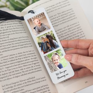 A high-quality metal bookmark with a black tassel rests on an open book. Three custom photos are printed vertically on the glossy white aluminium. The photos show a happy family, and a personalised message reads, "We love you Mummy xxx." The item is a perfect gift for Mother's Day, birthdays, or any special occasion. It is a heartfelt, personalised keepsake for a book lover.
