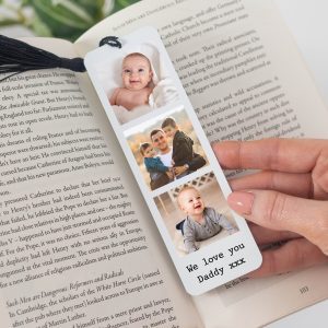Personalised photo bookmark featuring a collage of three family photos: a smiling baby, a father with two sons, and another photo of the same happy baby. The white metal bookmark has a black tassel and custom text that reads "We love you Daddy xxx". The bookmark is held over an open book, serving as a placeholder. A unique gift for Father's Day, birthdays, or as a keepsake for him.