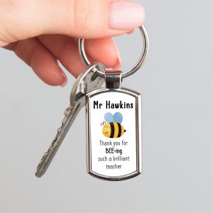 A high-quality metal keyring with a silver finish is held by a hand with light pink manicured nails. The keyring's rectangular pendant features a smiling cartoon bee with blue wings and black and yellow stripes. Above the bee, the customisable text "Mr Hawkins" is printed in black, and below, the text "Thank you for BEE-ing such a brilliant teacher" is also in black. The keychain ring and a single key are attached to the pendant.