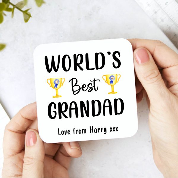 Wooden coaster held in two hands with the text "WORLD'S Best GRANDAD" printed on it. The text is in a mix of bold and script fonts, and there are two gold trophies with blue ribbons flanking the middle line. Below the main text, it says "Love from Harry xxx". This is a personalised gift for a grandad, perfect for Father's Day, birthdays, or as a thoughtful present from a grandchild. The coaster is square with rounded corners.