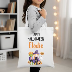 A young girl holding a personalised Halloween tote bag featuring a cute witch, monster, and ghost, with the text 'Happy Halloween' and the name 'Elodie'. The white canvas bag is a perfect trick or treat accessory for a child.