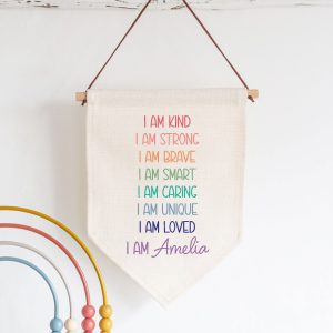 A beautiful, personalised linen flag with a positive affirmation for a girl. The fabric wall hanging sign features the phrase 'I AM KIND, I AM STRONG, I AM BRAVE, I AM SMART, I AM CARING, I AM UNIQUE, I AM LOVEDin a rainbow of pastel colours with a custom name at the bottom, perfect for a bedroom. Perfect gift for a girl, daughter, niece or granddaughter on their birthday or when starting school.
