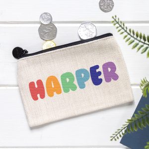 A personalised linen coin purse for kids with the customisable name "HARPER" printed in colourful rainbow letters. The small money pouch has a black zip and is shown on a white wooden surface with UK coins scattered around it. A green plant is visible in the corner. Perfect for storing birthday gift money for a son, daughter, niece or nephew.