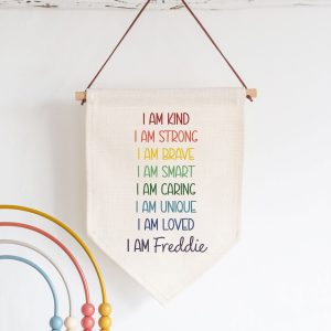 Personalised kids' linen flag with a positive affirmation rainbow design. The flag, made from thick, cream linen, has a wooden dowel and faux suede cord for hanging. It's personalised with the customisable name Freddie in a blue font. Perfect birthday or starting school gift for a son, boy, grandson or nephew.