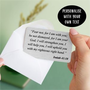 A hand holds a brushed silver aluminium wallet card, personalised with the bible verse "Fear not, for I am with you; be not dismayed, for I am your God: I will strengthen you, I will help you, I will uphold you with my righteous right hand. Isaiah 41:10." The card is partially slipped into a white envelope, with a black oval stating "PERSONALISE WITH YOUR OWN TEXT" in the top right corner, offering a clear call to action for this faith-inspired gift.