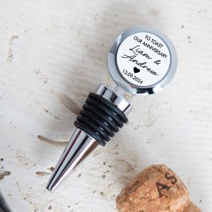 Personalised metal wine bottle stopper featuring a white top with the custom text "TO TOAST OUR ANNIVERSARY," customisable names "Liam & Andrew," and customisable date "12.09.2024" in black text. A small black heart is centred below the names. The shiny silver metal stopper has black rubber rings and a cone shape. It is lying on a light-coloured, textured surface next to a cork. The anniversary gift is ideal for a husband, wife, or couple.