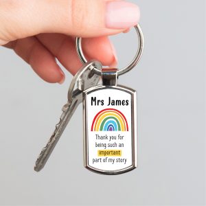A person holds a shiny, rectangular metal keyring. The keyring has a personalised insert with the customisable name "Mrs James" at the top. Below the name is a colorful rainbow arc. The bottom text says, "Thank you for being such an important part of my story," with the word "important" highlighted in yellow. The keyring is attached to a set of house keys.